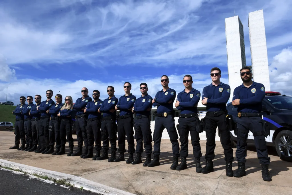 Fileira de policiais da Polícia Legislativa Federal em uniforme operacional azul marinho, posicionados em frente ao Congresso Nacional e viatura oficial.